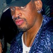 Black man in a baseball cap with lots of facial rings in lips, nose and ears wearing a blue shirt