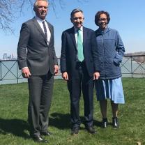 Three people standing outside on the grass with  body of water behind them, a tall white man in a suit with gray hair, and shorter white man in a suit and a black woman with glasses