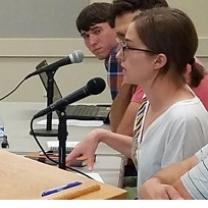 Young white woman with glasses and brown hair tied back speaking and gesturing at a podium with two guys at the table behind her 