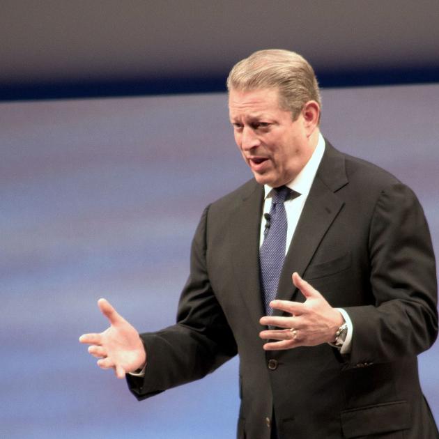 White man with graying dark hair in a suit holding his arms out in emphasis as he talks