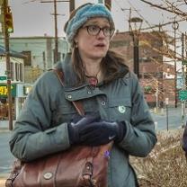 Young woman with brown rimmed glasses, long brown hair in winter clothes outside with her purse over her shoulder an holding it close to her body, looking like she is talking